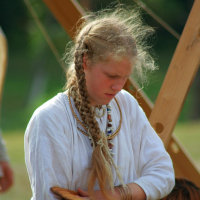 A young person with long, braided hair wears historical clothing and jewelry, including beaded necklaces and bracelets, while holding a wooden bowl. They appear to be participating in Semigallian Days '08, an outdoor historical reenactment or event, with a green, blurry background and wooden structures around them.