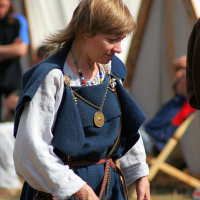 A person with short blond hair is dressed in medieval clothing, featuring a layered blue tunic and white undershirt, accessorized with a leather belt and pouch, necklaces, and a circular pendant. They are outdoors at Semigallian Days '08, with blurred tents and people in the background from the historical reenactment event.