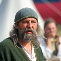 A man with long gray hair and a full beard, wearing a gray cap and green tunic with a white undershirt. He has a necklace with a metal pendant. The background shows a blurred figure of another person and a conical tent with a red structure partially visible at the Semigallian Festival, part of the 2008 events.