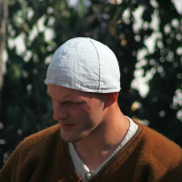 A person wearing a white cap and a brown tunic with a tassel stands outdoors at the 2008 Semigallian Days festival. The individual has a focused expression, and the background is filled with blurred greenery. The tunic appears to be made of coarse fabric, typical of historical reenactment clothing.