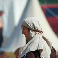 A person is dressed in medieval-style clothing, featuring a light headscarf and a dark tunic with a white undershirt. The background shows a blurred outdoor setting with a conical tent and people who appear to be part of the 2008 Semigallian Days historical reenactment or festival.