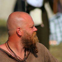 A bald man with a thick red beard is dressed in a brown tunic and adorned with necklaces, including one with a large pendant. He looks to his left, likely reminiscing about Semigallian days. The background is out of focus, showing another person holding an object as sunlight illuminates his face and shoulders.