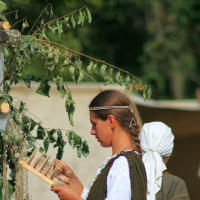 A woman dressed in traditional Semigallian attire, with a white blouse and dark vest, stands outdoors focused on weaving or crafting with a wooden tool during the 2008 Semigallian Days. Her hair is tied back in a ponytail. Another person, partially visible, is standing nearby wearing a white headscarf. Trees and greenery are in the background.
