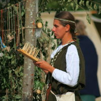 A woman dressed in medieval-style clothing is seen weaving or inspecting threads on a wooden loom at the Semigallian Days festival. She has long hair tied back with a headband, wearing a white undergarment and dark overdress, with a pouch at her waist. Behind her are hanging necklaces and greenery, part of the 2008 historical reenactment setup.