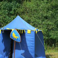 A blue medieval-style tent with yellow trim stands on grassy ground. The tent has an opening in the front, revealing a dark interior. Beside the entrance is a blue shield featuring a yellow griffin, reminiscent of the days gone by. The background consists of dense green foliage.