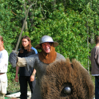 A person dressed in medieval armor, including a chainmail shirt and a metal helmet from the 2008 Semigallian Days, is smiling and wielding a sword in one hand while holding a fur-covered shield in the other. They stand outdoors with trees in the background and other people dressed in historical clothing nearby.