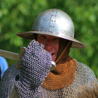 A person in medieval armor, including a chainmail suit and a metal helmet, holds a large axe during the Semigallian Days festival. The silver helmet with rivets gleams under the sun, while their protective suit covers their neck with rusty colored material underneath. Blurred greenery forms the backdrop.