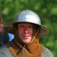 A person dressed in medieval armor, including a metal helmet and chainmail, looks directly at the camera. The individual has a faint smile and the background is blurred greenery, suggesting an outdoor setting during Semigallian days 2008. The chainmail hood is brown, indicating age or a specific design choice.
