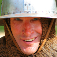 A person is smiling and wearing Semigallian medieval reenactment armor, which includes a metal helmet with rivets and a chainmail coif covering the head and neck. The background is blurry, emphasizing the individual’s detailed facial expression and historical attire. The lighting is bright and natural.