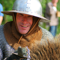 A person dressed in medieval chainmail armor and a metal helmet, smiling at the camera during Semigallian Days 2008. They have a chainmail coif under the helmet and are holding a fur-covered shield. The background is blurred, showing greenery and someone in modern clothing.