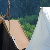 Two canvas tents are pitched in an open grassy area with a backdrop of dense, green trees, reminiscent of Semigallian Days. One tent is beige and constructed in a traditional triangular shape with a small opening. The other tent is white and has a larger and more structured appearance, evoking the spirit of the 2008 event. Various camping items are scattered around.