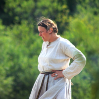 A person is shown standing outdoors during the '08 Semigallian days festival, wearing a long, white garment with a brown belt. The individual has short, light-colored hair and appears to be looking down or to the side. The background is a blurred view of green foliage, suggesting a natural, outdoor setting.
