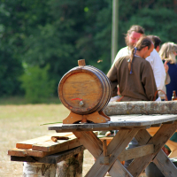 A small wooden barrel is placed on a rustic wooden table, set outdoors with a background of green trees. Several people are gathered near the table, engaged in conversation. A wooden bench and scattered bottles are visible in the foreground, evoking memories of relaxed Semigallian days and casual outdoor gatherings.