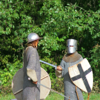 Two people dressed in medieval armor engage in a mock sword fight in a wooded area during the annual Semigallian Days event. They wear chainmail tunics and helmets. One holds a round shield, and the other has a shield with a black cross. Both hold swords, with a backpack on the ground behind them.