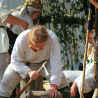 A person dressed in historical Semigallian clothing is chiseling wood while kneeling on the ground. Other individuals, also in period attire, are visible in the background. The setting appears to be a days-long outdoor reenactment camp with greenery and rustic wooden structures, reminiscent of 2008 events.