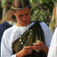 A woman with long brown hair, adorned in a simple headband, wearing a white blouse and a dark green draped garment, stands outdoors. She is focused on weaving or knotting a bundle of fibers in her hands during Semigallian Days 2008. She also wears a necklace made of light-colored beads. The background is blurred greenery.