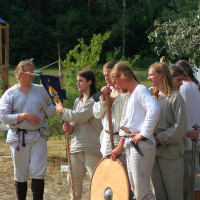 In a 2008 reenactment, a group of people dressed in historical medieval attire stands outdoors in a forest clearing. They are wearing white tunics and holding various props like wooden poles, a round shield, and a flag. One person appears to be speaking to the group. Trees and greenery are visible in the background, reminiscent of old Semigallian days.