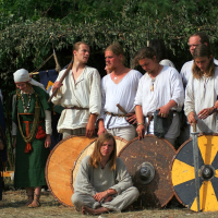 A group of people, dressed in historical Viking attire, pose outdoors as if transported from Semigallian days. Men and women stand behind three large round shields with colorful designs, and a young man sits on the ground in front. The background is lush green foliage and a structure made of branches. Some wear jewelry and ornate clothing.