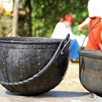 A large, black, cast-iron kettle with a handle and a stir stick rests on a wooden surface outdoors. In the blurred background, two people dressed in historical attire, including a white shirt and a red skirt, are engaged in conversation. This scene is from the Semigallian festival held in 2008, surrounded by lush greenery.