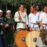 A group of people dressed in medieval attire are gathered together at a Semigallian festival. On the left, a woman wears a white headpiece and elaborate jewelry, while the men around her wear simple tunics. Some men hold round wooden shields and swords. The background features greenery and a rustic setup reminiscent of 2008 Semigallian days.