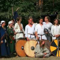 A group of eleven people in medieval clothing stands outdoors during the Semigallian Days. Some wear white tunics while others are dressed in blue and green robes adorned with jewelry. Three hold round wooden shields, and one person kneels in front with blonde hair. The background has green foliage and trees.