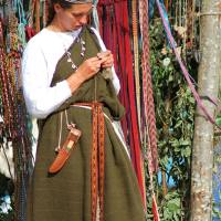 A woman wearing a medieval-style green dress and a white long-sleeve shirt is standing in front of an array of colorful ribbons hanging in the background. She is focused on her hands, possibly crafting or tying a knot. A brown belt with a sheathed knife hangs at her waist, reminiscent of 2008's Semigallian Days celebration.