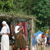 Three people dressed in historical clothing are partaking in an outdoor, medieval reenactment at Semigallian Days '08. One person stands under a canopy with colorful ribbons; another crafts an item on the right, while a third, seen from the back, is to the left. Trees and a tent are visible in the background.