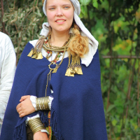 A person dressed in traditional attire stands outdoors during Semigallian Days 2008. They are wearing a blue cloak, several necklaces with large pendants, and arm bracelets. A white headscarf with a golden headdress adorns their head while green foliage is visible in the background.