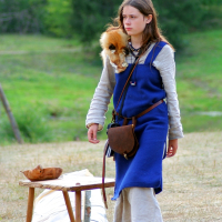 A young person wearing historical Semigallian attire, including a blue tunic over a beige shirt and beige pants, stands outdoors on a dry grassy area reminiscent of days past. They have a brown leather bag and a small fur accessory. A simple wooden table and bench with wooden bowls and a spoon are nearby, with green trees in the background.