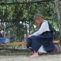 A woman and a child dressed in traditional medieval clothes sit on a wooden bench under a canopy of leafy branches. The woman is occupied with a task, while the child is engrossed in another activity. This rustic, natural setting harks back to the ambiance of Semigallian Days '08, surrounded by greenery.