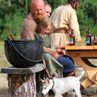 A bearded man sits on a log bench with a young child on his lap near a large black pot, reminiscent of the lively 2008 event, Semigallian Days. Next to them are two men seated at a wooden picnic table with bottles. A small white puppy on a leash stands in the foreground of the outdoor, wooded area.