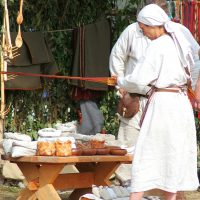 At Semigallian Days 2008, a person in a white tunic and headscarf, wielding a weaving tool with red threads, stands near a wooden table filled with baskets and bowls. Behind them, hanging fabric and wooden utensils adorn the structure, evoking an outdoor market or fair.