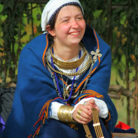 A smiling woman wearing a white headscarf and a royal blue cloak adorned with colorful tassels sits in front of a greenery backdrop. She wears various necklaces and bracelets, holding a small wooden instrument in her hands, reminiscent of the vibrant Semigallian days. She appears relaxed and content.