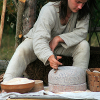 A man with long brown hair, dressed in a light-colored medieval-style tunic and pants, uses a small hand tool to grind grain on a stone mill. Surrounding him are wooden bowls filled with grain and flour, and a woven basket. The scene evokes the historical charm of Semigallian days circa 2008 amidst outdoor greenery.