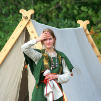 During the 2008 Semigallian Days, a woman stands in front of a canvas tent, wearing a green dress adorned with various accessories. She has her right hand raised to her forehead, partially covering her eyes. The background features green foliage, suggesting an outdoor setting. She holds a white cloth with red edges in her left hand.