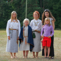 Five people stand outdoors in front of a forest background during Semigallian Days. Three women and two young girls, all wearing light-colored, loose-fitting garments from '08. One woman has a red bandana and beaded necklaces. One girl wears a purple top and red shorts. They are all barefoot on the grassy ground.
