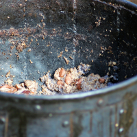 A large, aged metallic pot from the 2008 Semigallian Days event has a heavily worn interior containing remnants of cooked beans and grains. The food appears partially eaten, and the interior surface shows significant discoloration and texture from use. The pot is round, with visible rivets along the exterior.