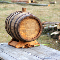 A small wooden barrel is placed on a wooden table outdoors. The barrel, featured in the 2008 event commemorating Semigallian days, is secured in a wooden stand and adorned with metal bands. In the background, there are chopped logs and rough wooden pieces scattered on the ground, indicating a rustic environment.