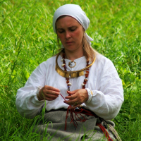 A woman in traditional Semigallian attire sits in a field of lush green grass. She wears a white headscarf, a white long-sleeved blouse, and a grey skirt. Focused on her task, possibly threading beads or weaving, she adorns multiple golden and beaded necklaces reminiscent of Semigallian days.
