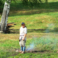 A person dressed in historical Semigallian clothing, including a tunic, belt, and leggings, stands on a grassy area. They are looking down at a spot with faint smoke visible. In the background, there is a stack of wood and a large tree. A wooden bucket and some utensils lie on the grass nearby.