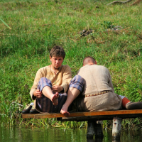 Amid the green grass and slightly sloped, natural landscape, two individuals clad in simple, weathered clothing sit on a wooden platform by the calm water's edge. The scene, reminiscent of Semigallian days past even in 2008, features one person tending to their feet while the other gazes into the serene waters.
