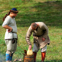 At the Semigallian festival, two people dressed in medieval-style clothing are in a grassy outdoor setting. One person, with long hair and a blue cap, stands with hands on hips, watching the other, who is bald and bent over, placing a round loaf of bread into a wooden barrel. They both wear tunics, trousers, and boots.