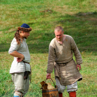 Two people wearing medieval-style clothing are standing on grassy terrain. One, with long hair and a headband, is watching the other, who is bald and holding a bucket by its wooden handle. Both wear neutral-toned tunics, belts, and colorful leg wraps amidst a green, sunlit backdrop reminiscent of Semigallian Days 2008.
