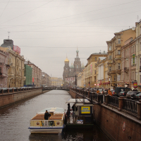 A boat cruises along a narrow canal in St. Petersburg, flanked by classic European architecture under a cloudy sky, with a bridge and the onion domes of a prominent church visible in the background