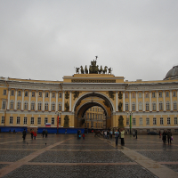 The general staff building arch glows warmly in yellow hues, while a horse-drawn chariot perches atop, overlooking a cloudy sky and a scattering of visitors in St. Petersburg's palace square.