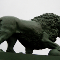 A majestic bronze lion statue, presented in profile, stands with a poised and muscular posture atop a stone plinth against a gray sky in St. Petersburg.