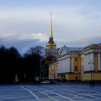 A quiet, expansive city square in St. Petersburg with crisscrossing white lines on the asphalt, leading to a historic building with a golden spire piercing a blue sky scattered with clouds.