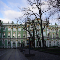 A baroque-style green and white palace in St. Petersburg, with ornate white sculptures and windows under a blue sky, flanked by bare trees, in a cobblestone-paved courtyard.