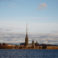 A golden spire pierces the sky above a historic fortress on a waterfront in St. Petersburg, under a vast expanse of clouds tinged by the soft light of either dawn or dusk.