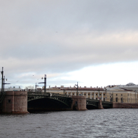 Overcast sky over a wide river with a classic multi-arched bridge leading to historic European-style buildings and a prominent tower in the background, capturing a serene yet somber urban landscape in St.